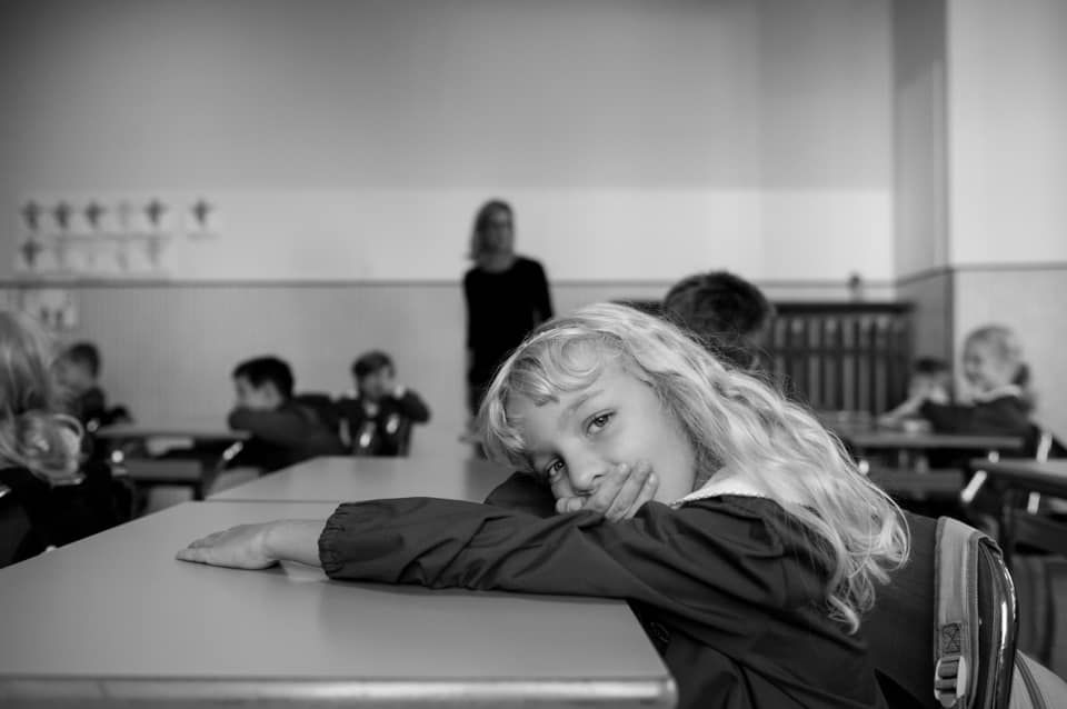 Black and white photo of a young blonde girl resting her head on a desk in a classroom full of students.