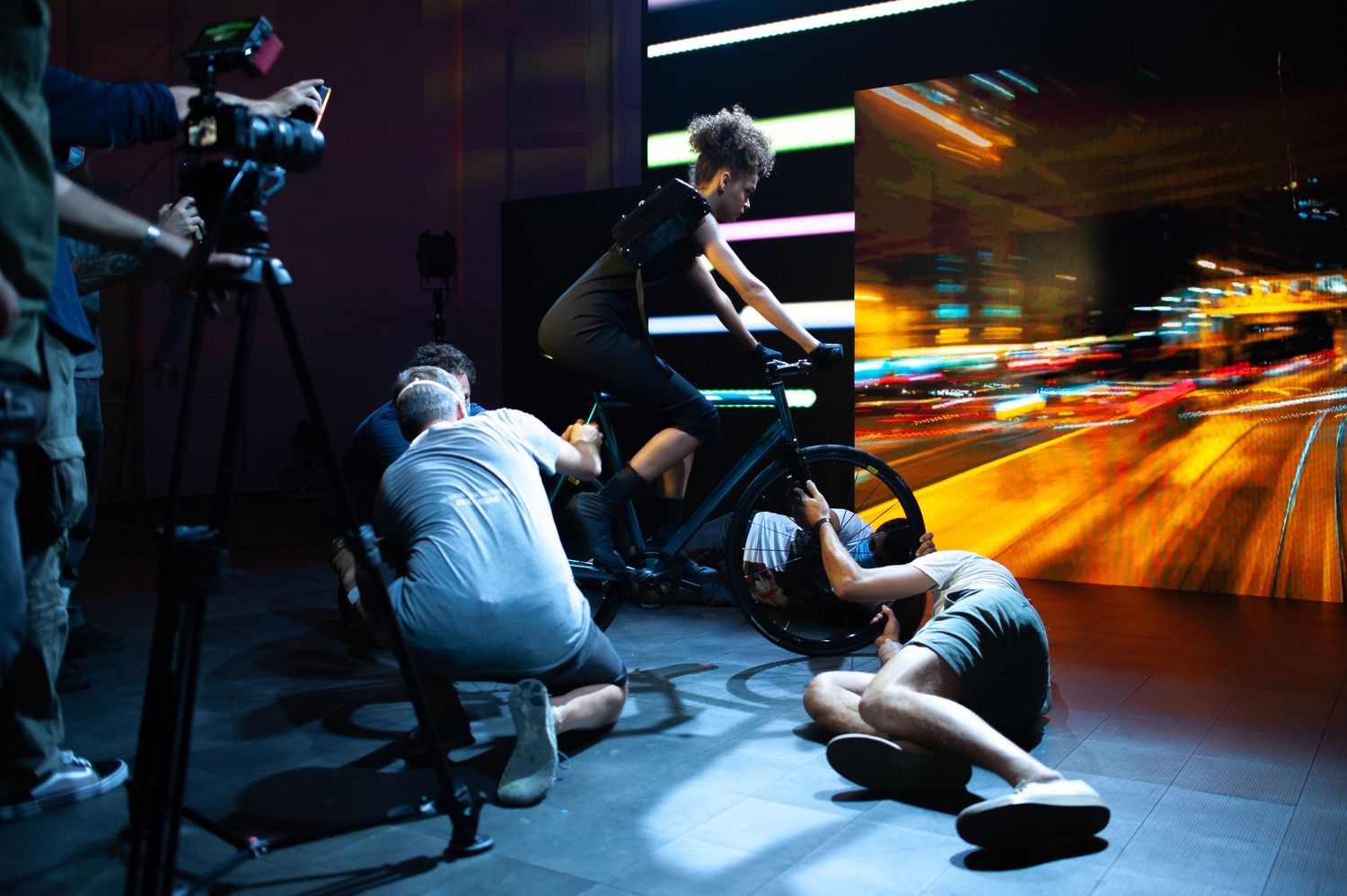 Film crew shooting a cyclist on a stationary bike against a vibrant motion-blur city backdrop in a studio.