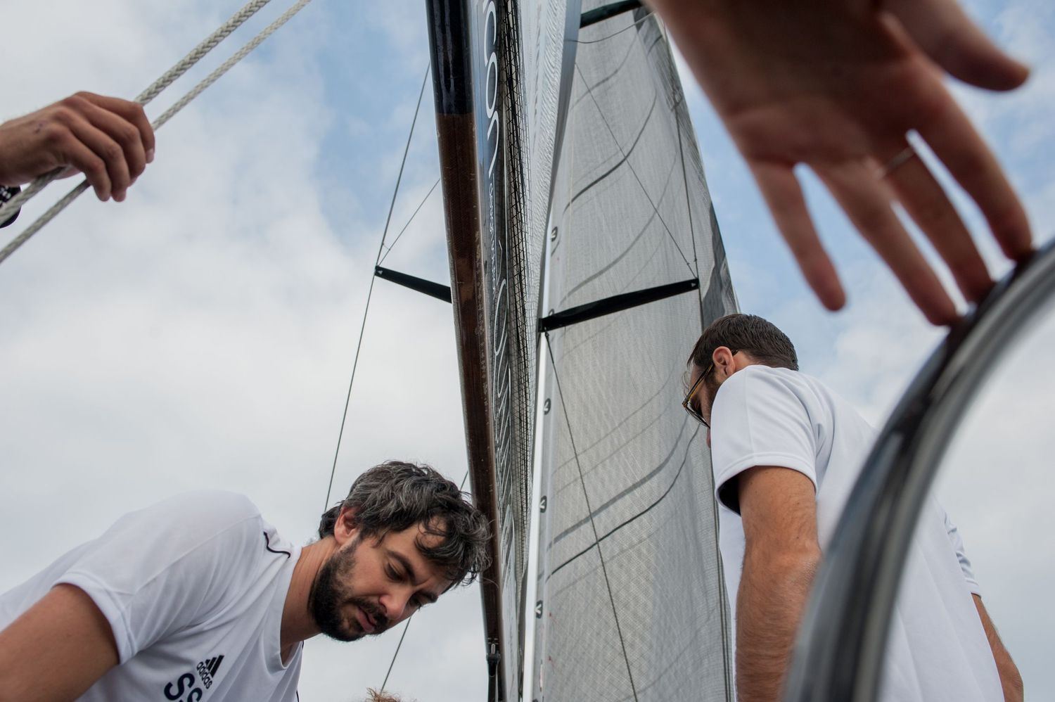 Two sailors in white shirts work on rigging aboard a sailboat with white sails against a cloudy sky.