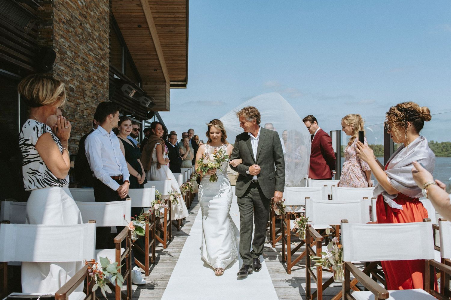 Outdoor wedding ceremony with guests seated on white chairs along aisle runner at rustic stone venue.
