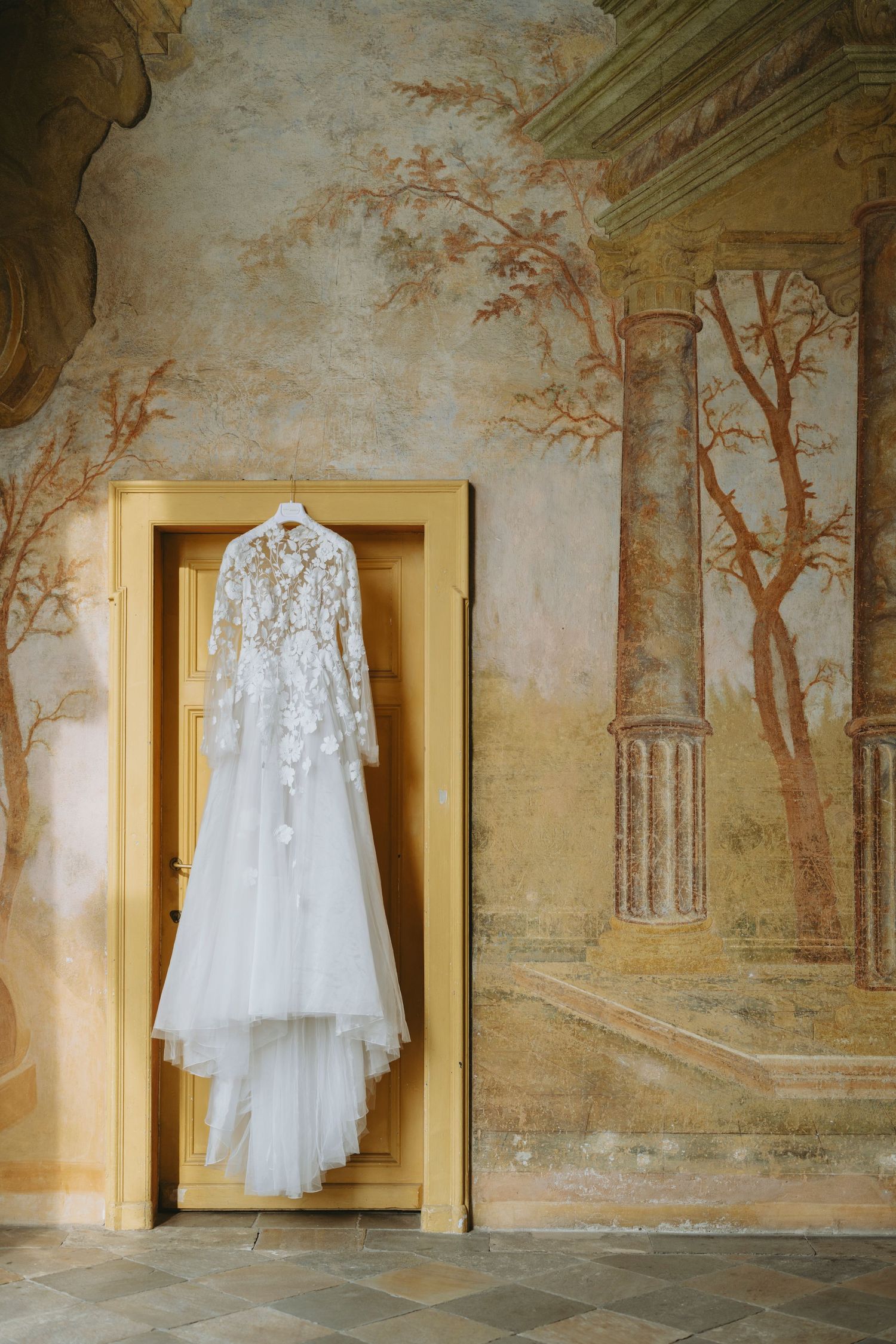 An elegant white lace wedding dress hangs on a door frame against a wall with classical mural artwork depicting trees.
