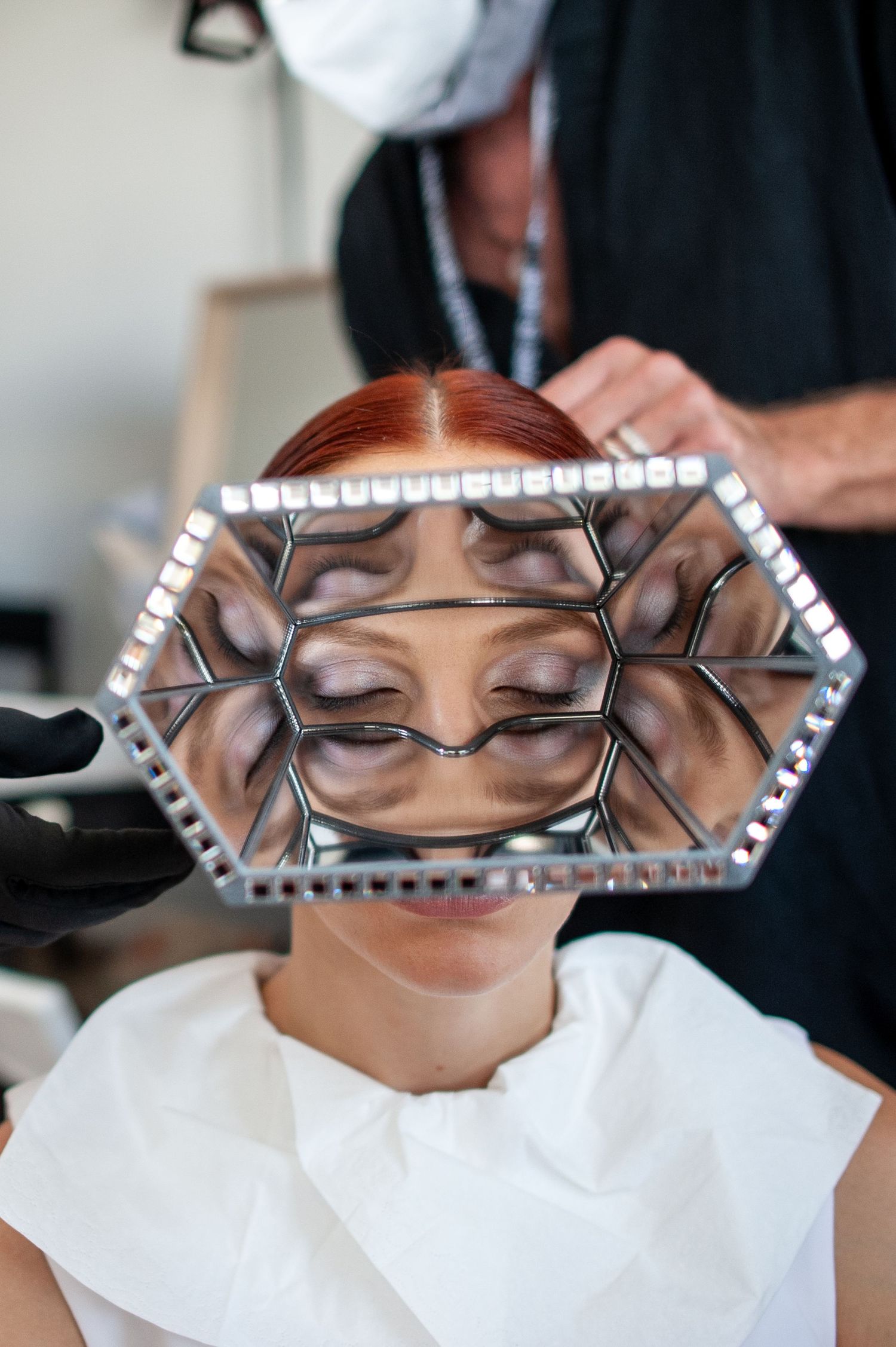 Hairstylist holding hexagonal mirror reflecting client's face with dramatic eye makeup in a salon.