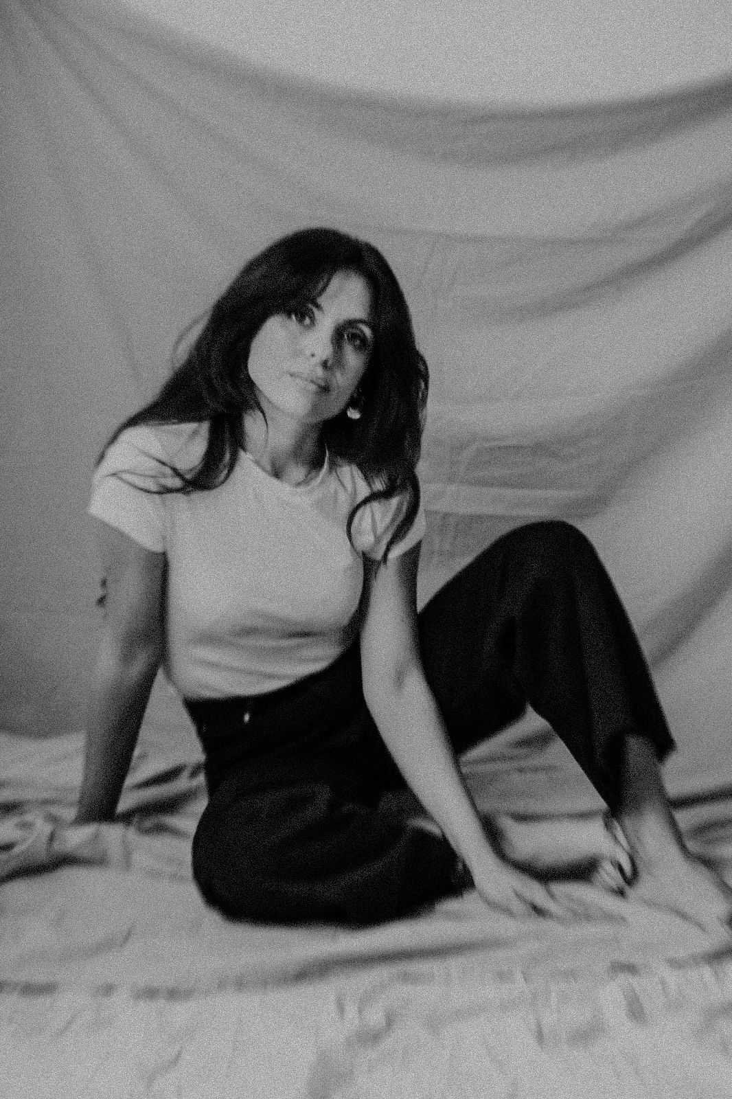 Black and white portrait of someone sitting on fabric wearing a white t-shirt and dark pants in a casual pose.