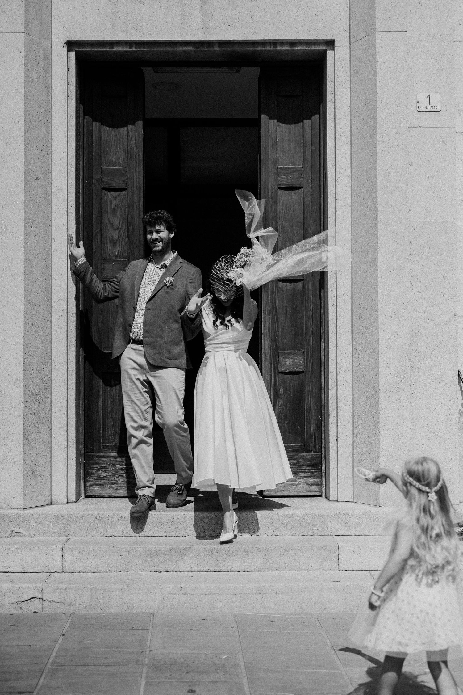 A black and white photograph shows a young child in a dress running across a building's porch or walkway.