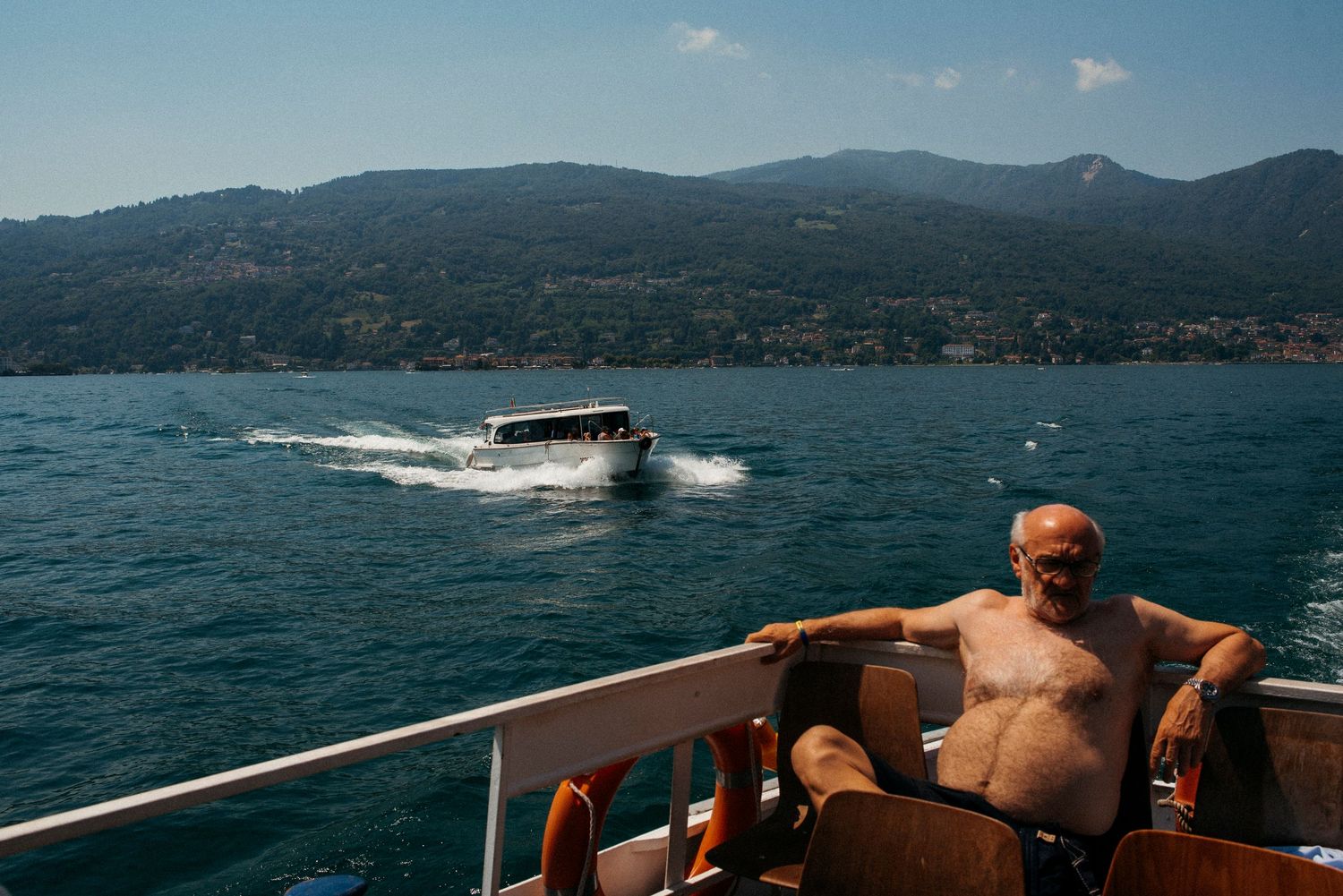 Shirtless man relaxing on boat deck chair with motorboat and mountain lake scenery in background on sunny day.