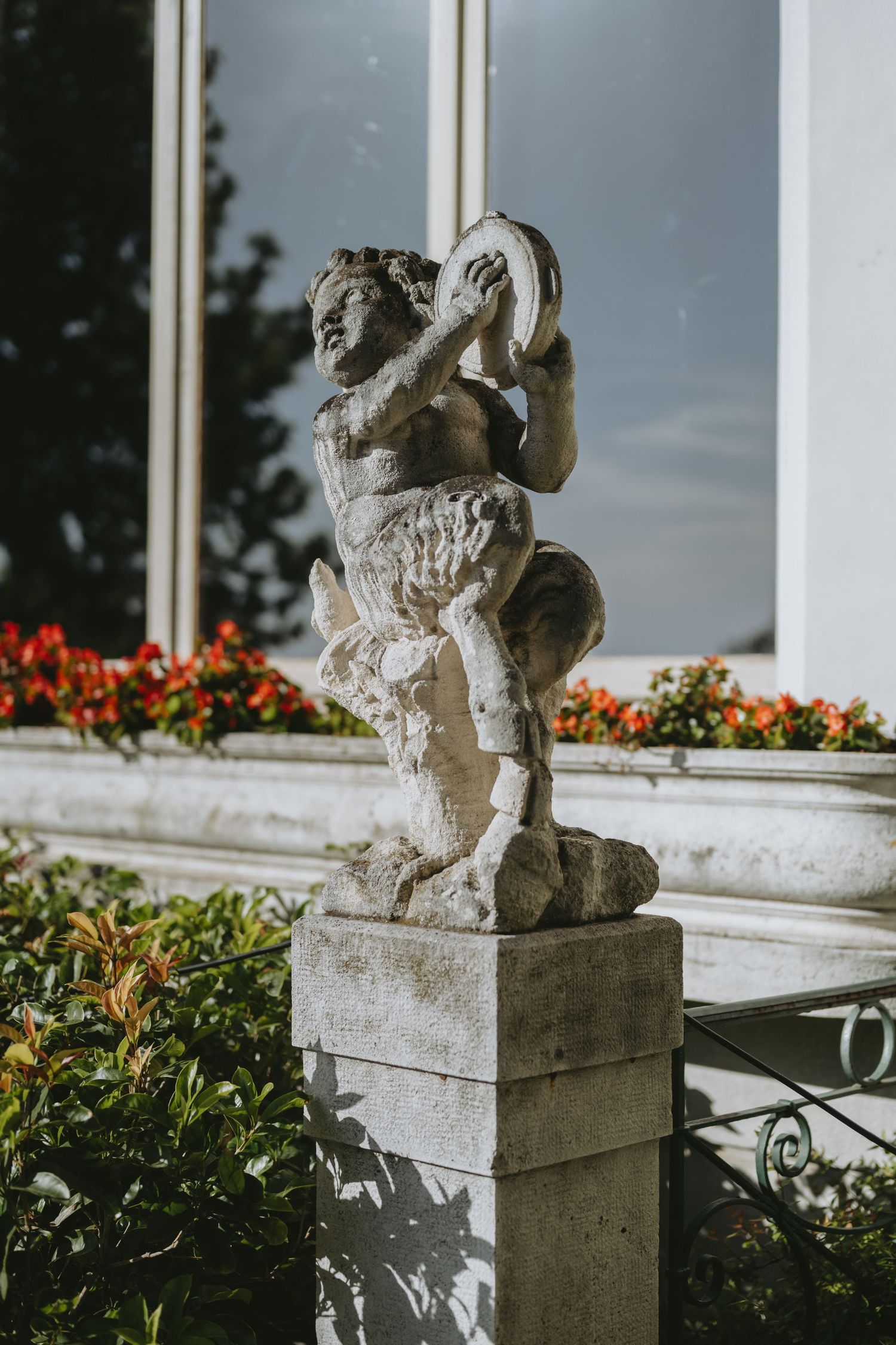 Weathered stone cherub statue holding a tambourine on a pedestal, surrounded by red flowers and greenery outside a white building.