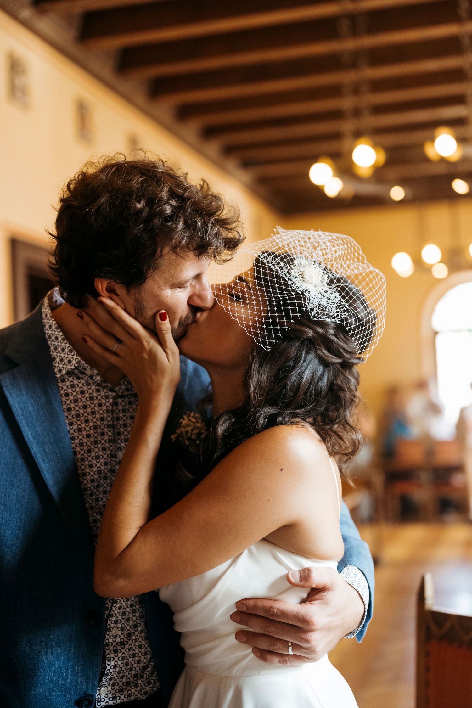 A romantic wedding kiss captured in a rustic venue with warm lighting and the bride wearing a vintage-style birdcage veil.