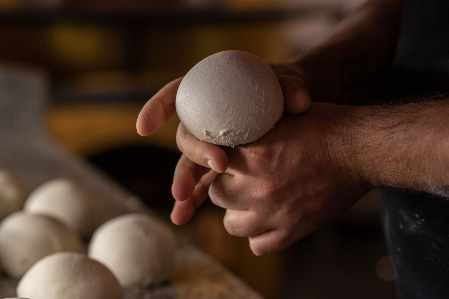 Baker's hands shaping a round bread dough ball with more dough portions visible on a floured surface in a bakery.