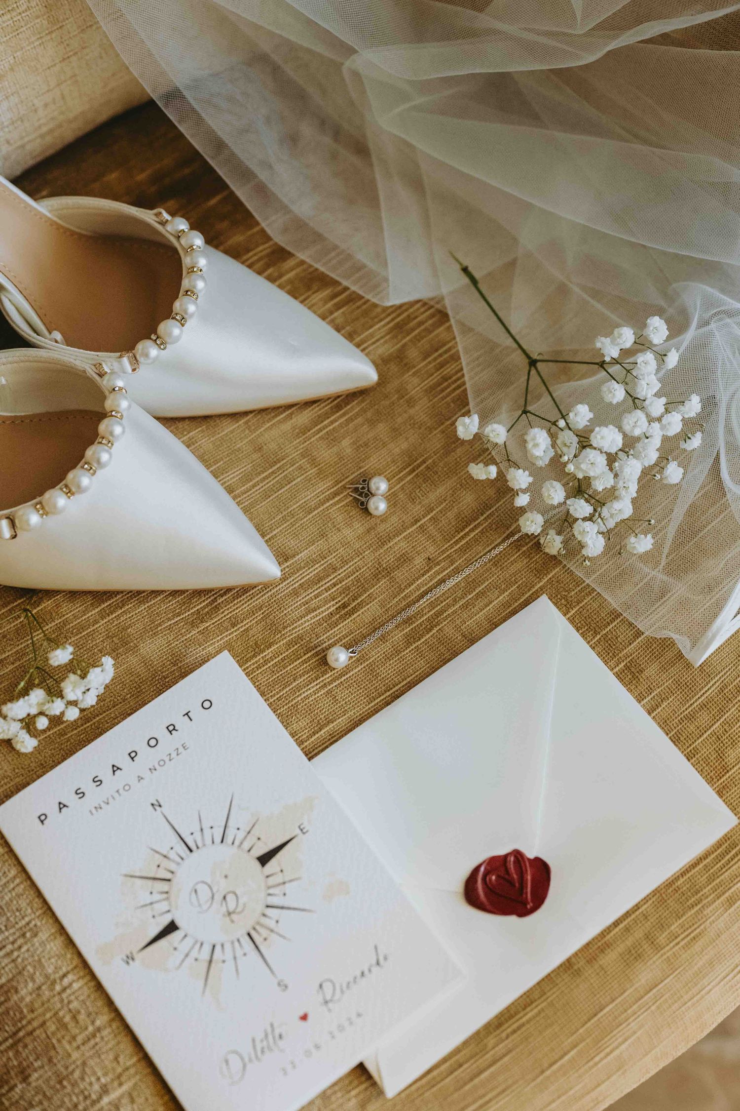 Elegant bridal flat lay with pearl heels, wax seal envelope, wedding invitation, baby's breath, and veil on wood surface.