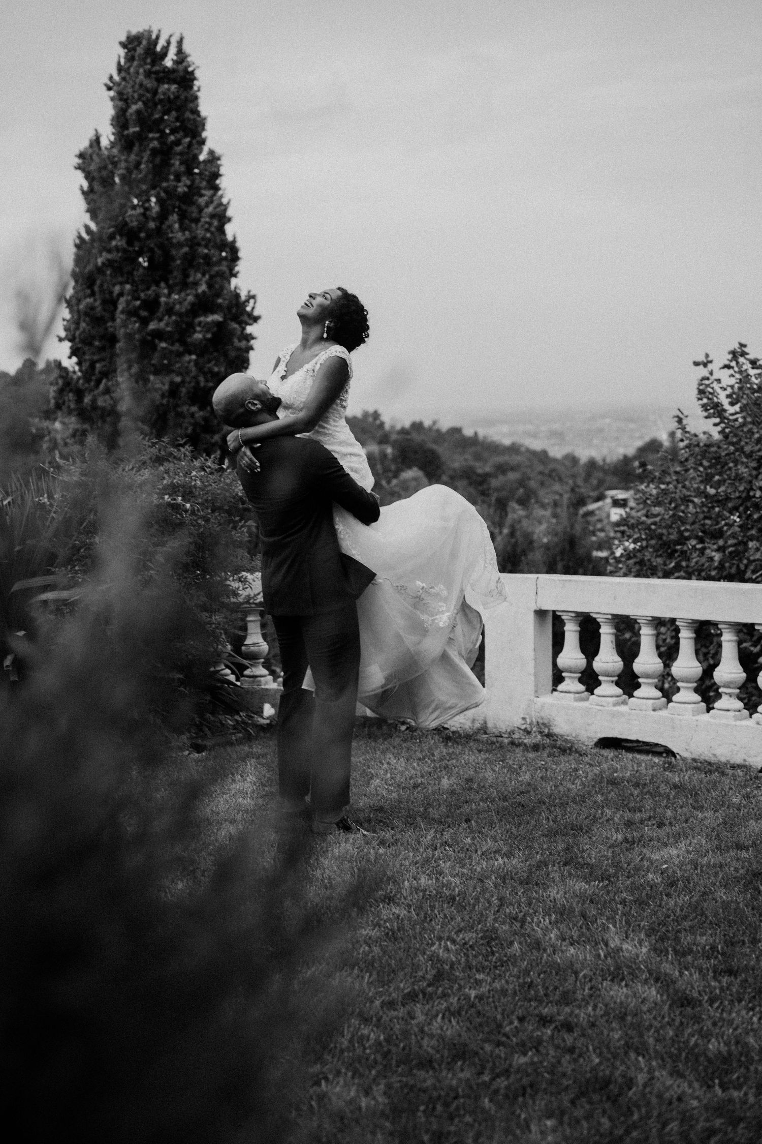 Groom lifting bride in white gown outdoors in black and white wedding photo with lush garden and balustrade.