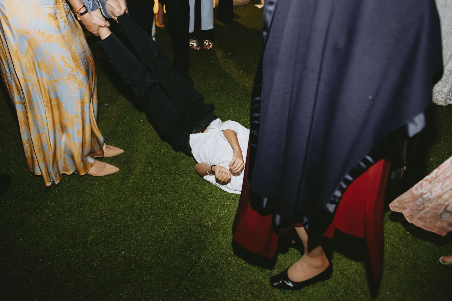 Child lying on green grass surrounded by adults in formal attire at an outdoor event.