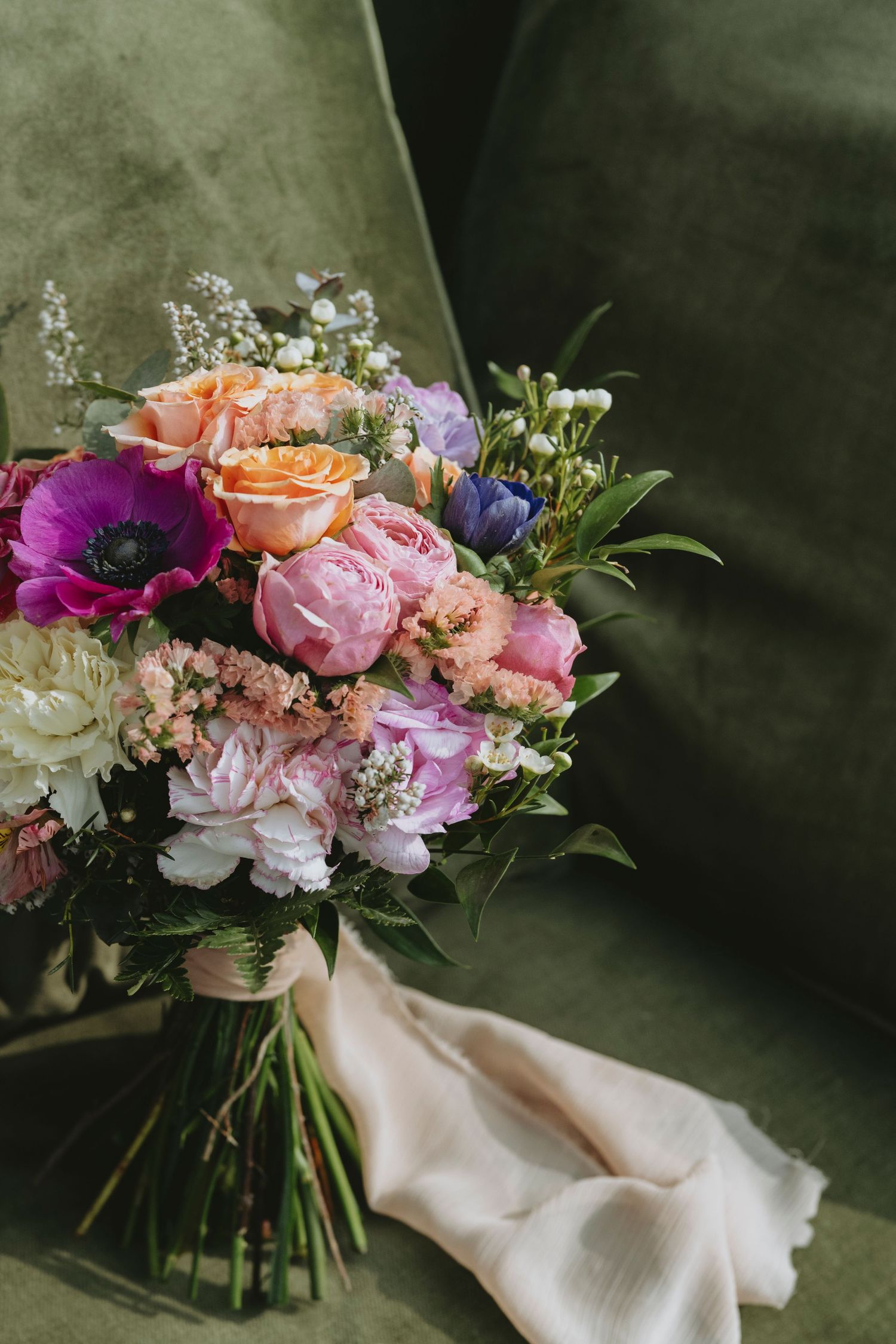 A lush wedding bouquet with pink peonies, peach roses, purple anemones and greenery arranged against a dark background.