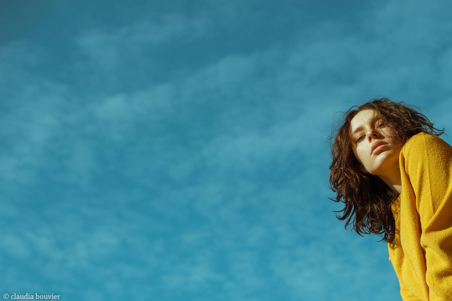 Young woman in yellow sweater gazing upward against a vibrant blue sky with soft clouds.