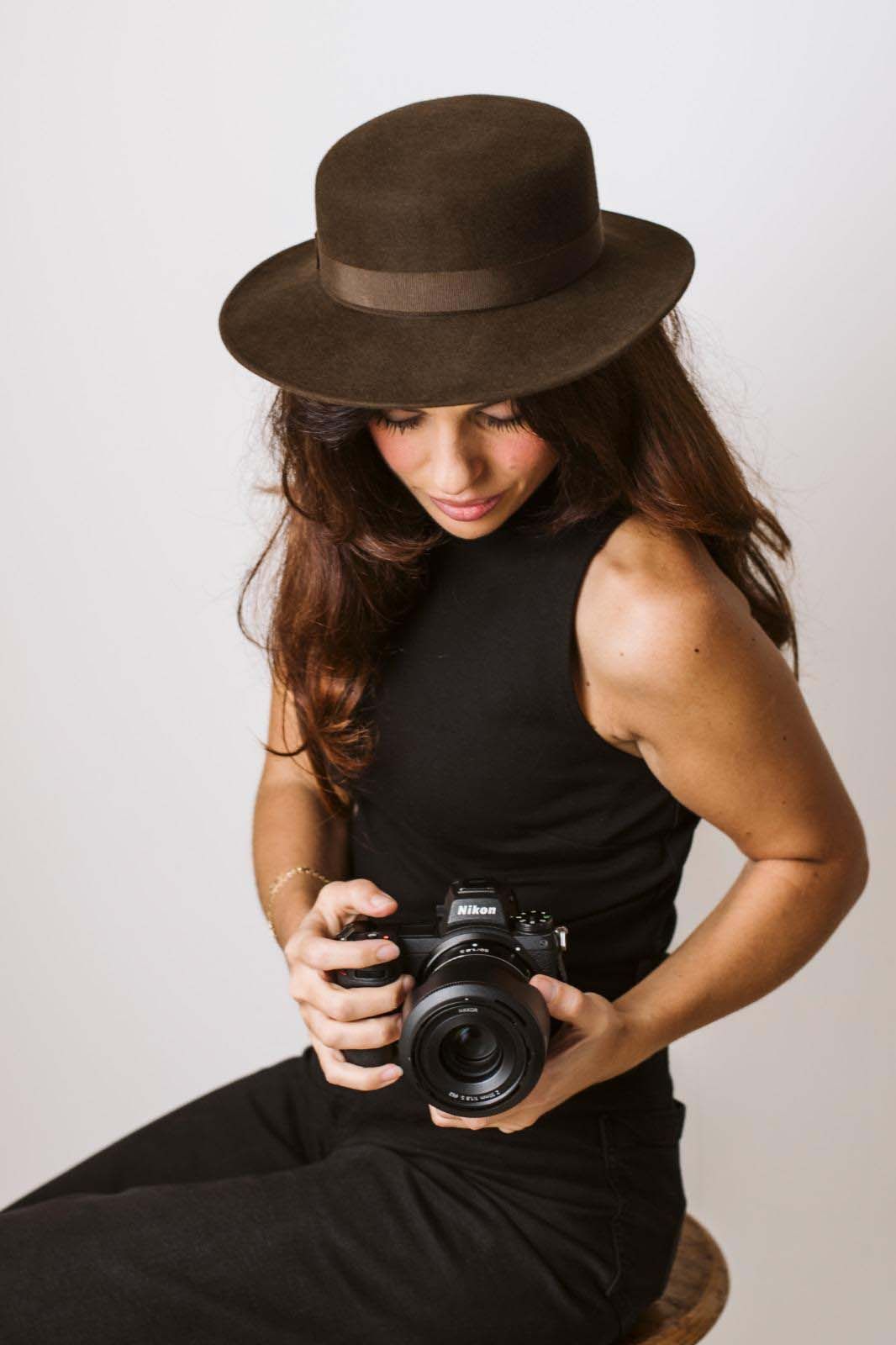 A photographer in a black dress and brown hat poses with a vintage camera against a white background.