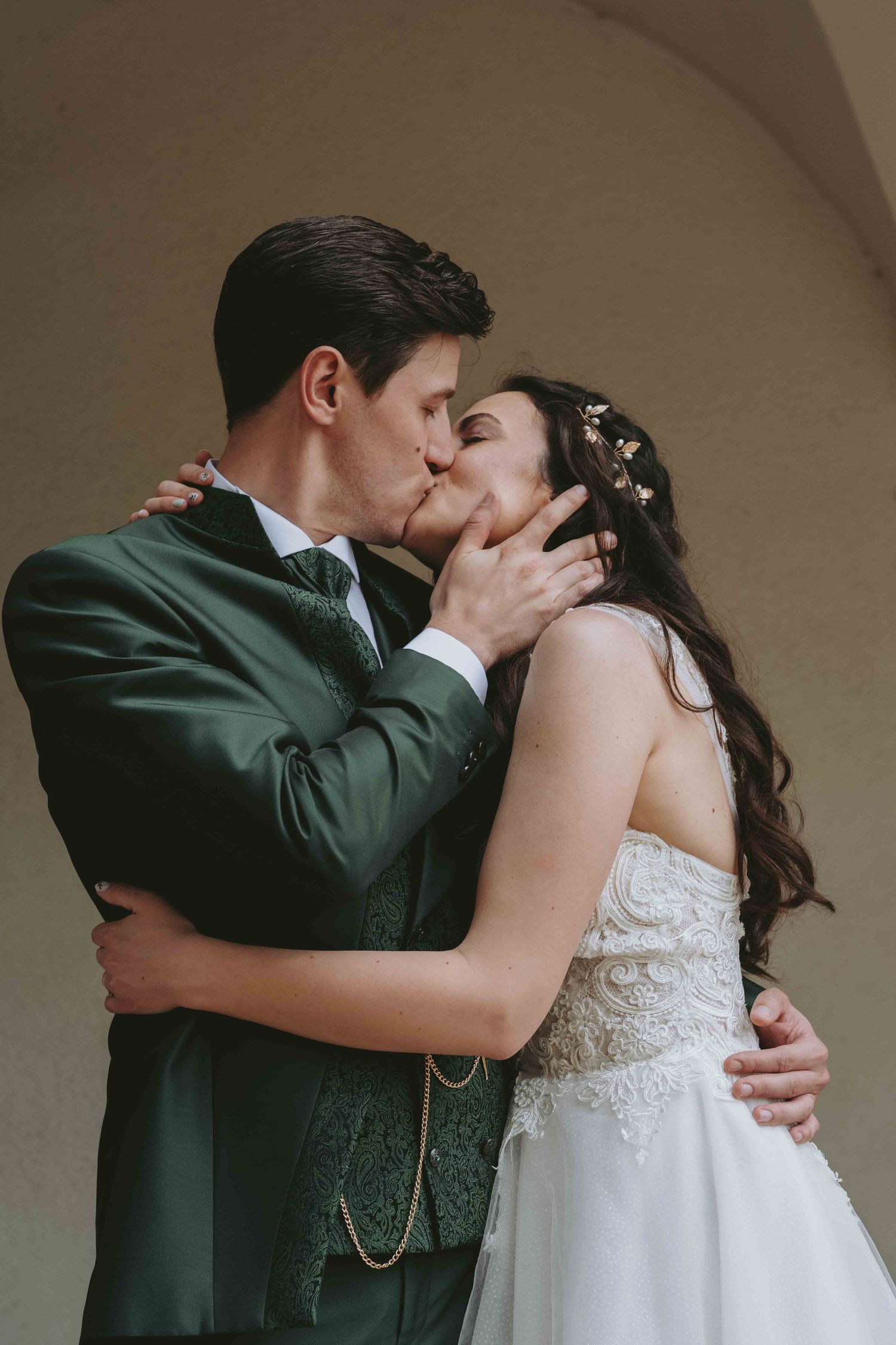 A bride and groom share a romantic first kiss, dressed in a white gown and dark green suit at their wedding.