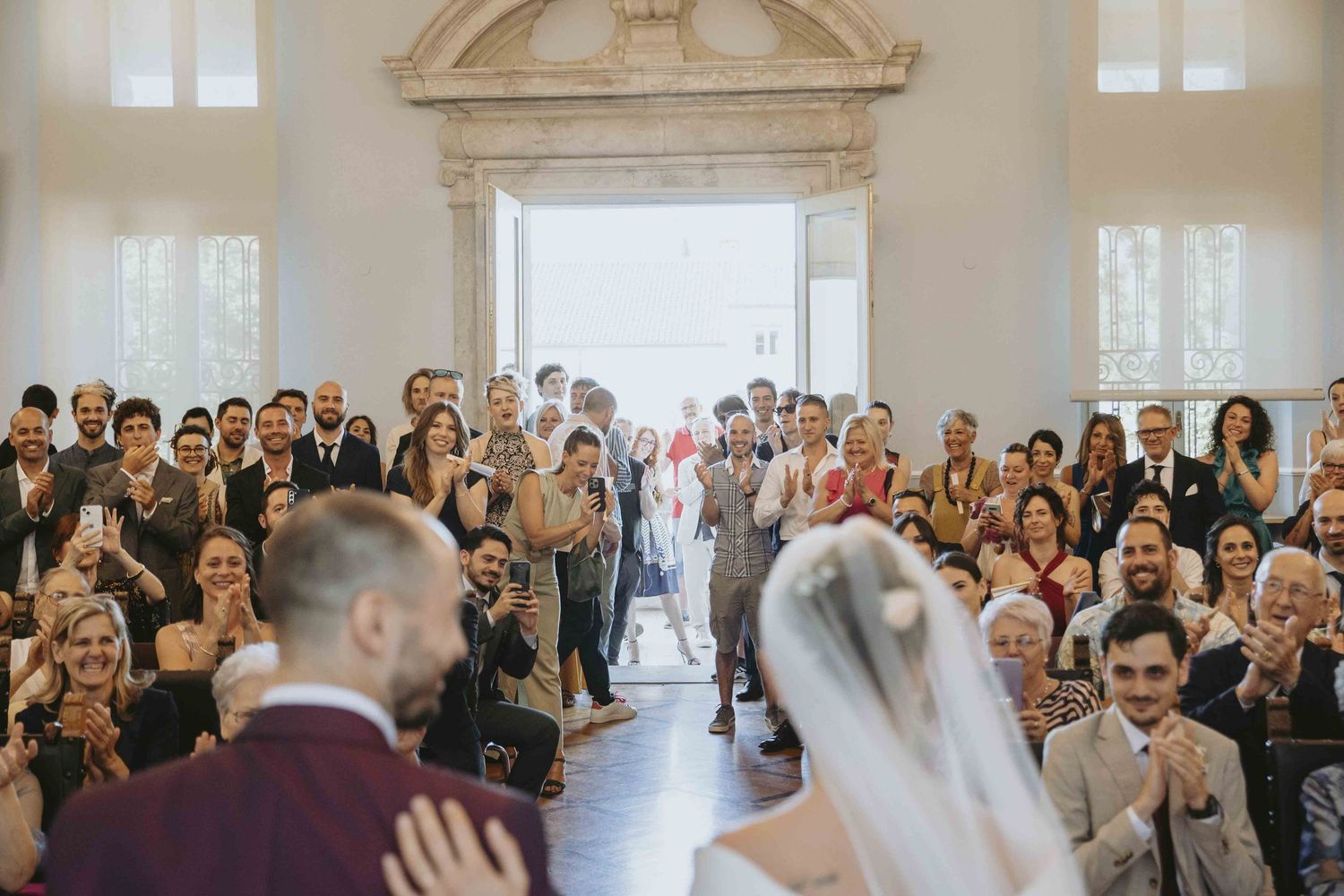 Wedding ceremony with bride and groom at altar, guests applauding in elegant venue with grand doorway.
