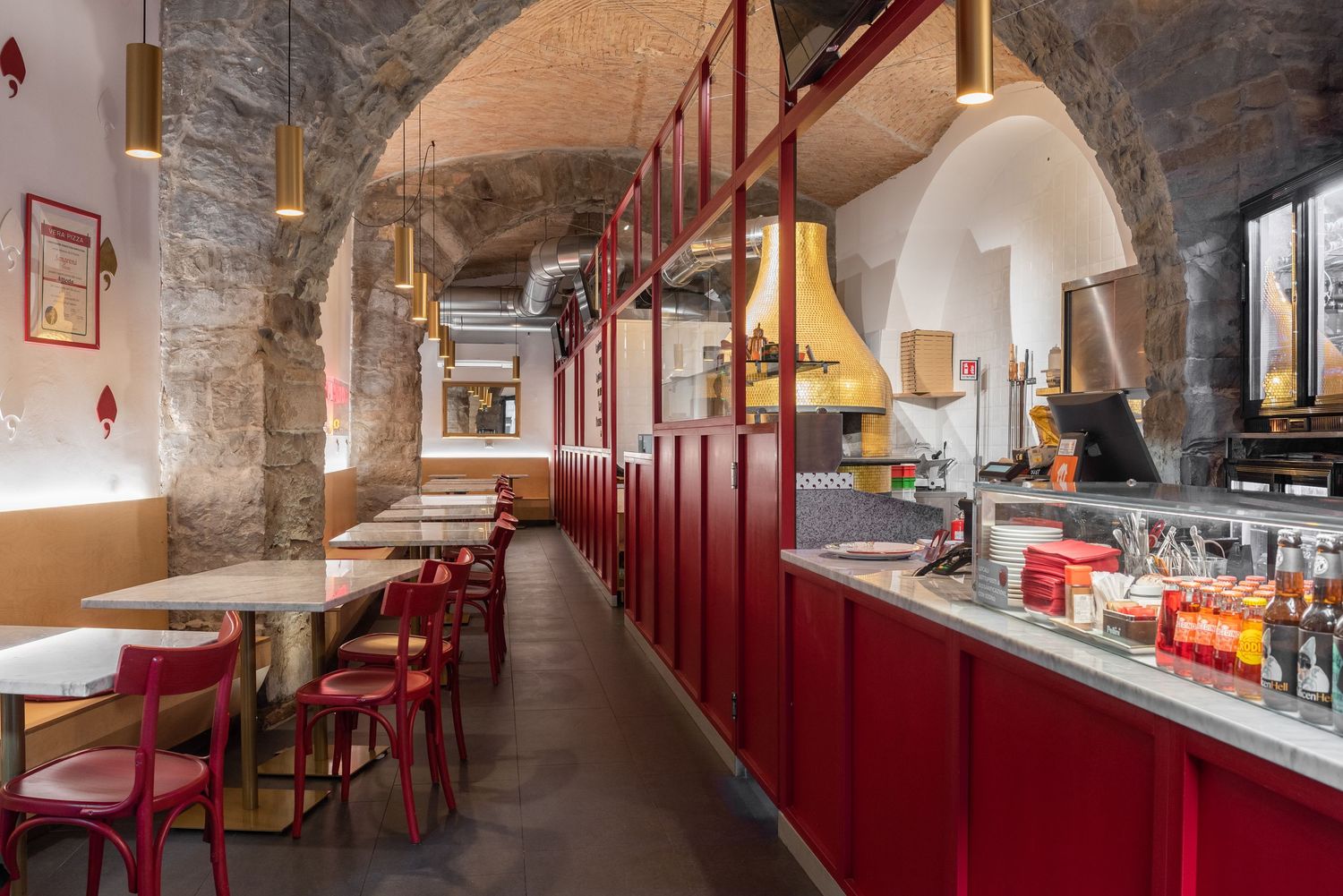 Cozy restaurant interior with red booths, stone arched walls, wooden tables, and a service counter under warm pendant lights.