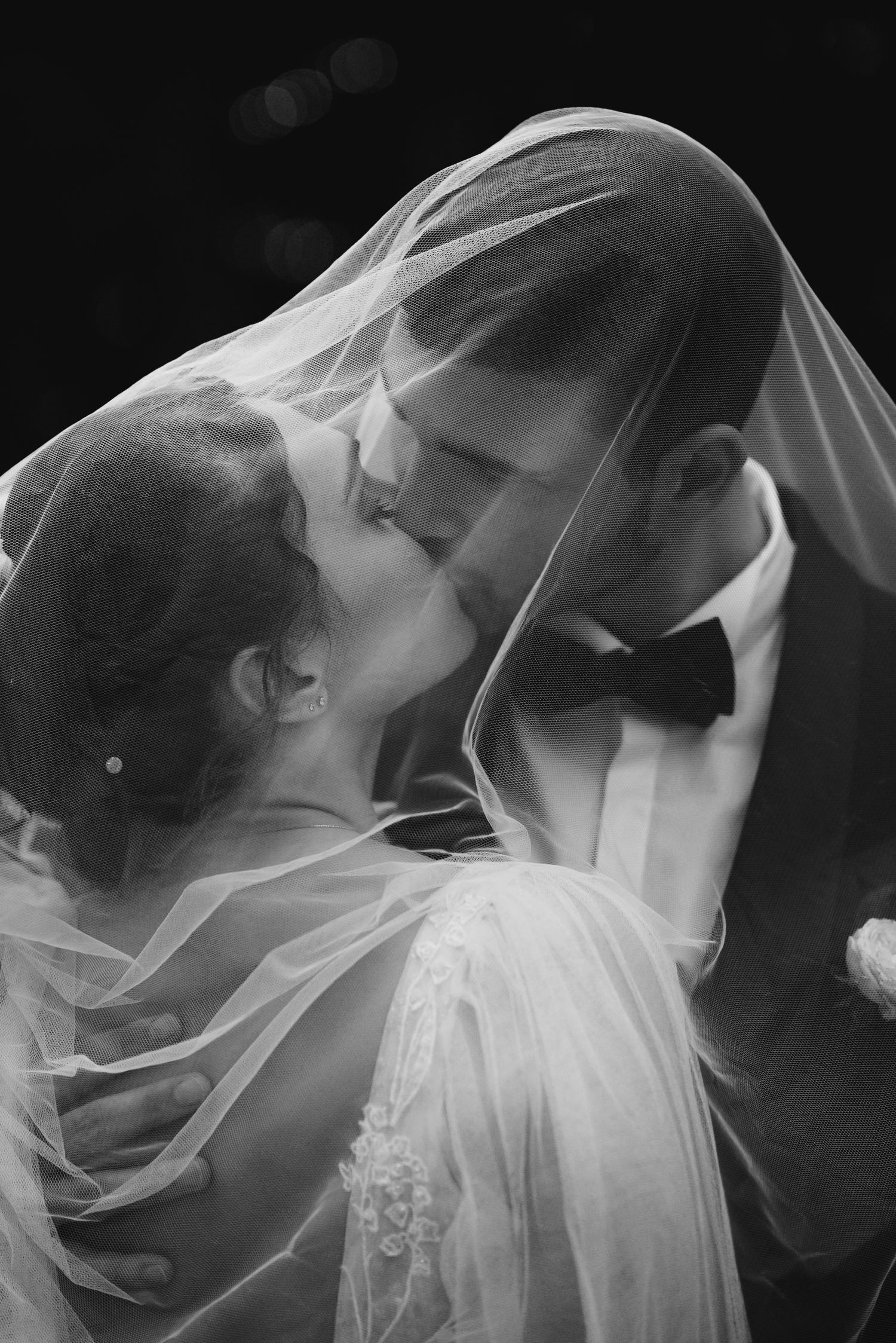 Black and white photo of bride and groom sharing a kiss under a flowing wedding veil.