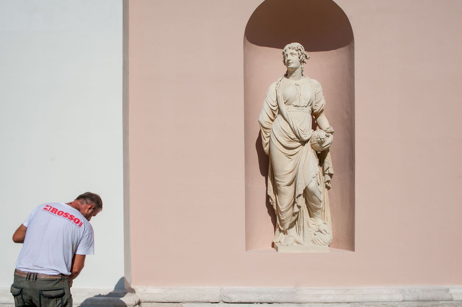 Man in white shirt stands beside a pink wall with a classical stone statue in an arched niche.