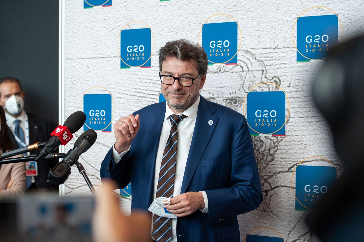 Politician speaks at G20 Italia 2021 press conference, standing before branded backdrop in blue suit.