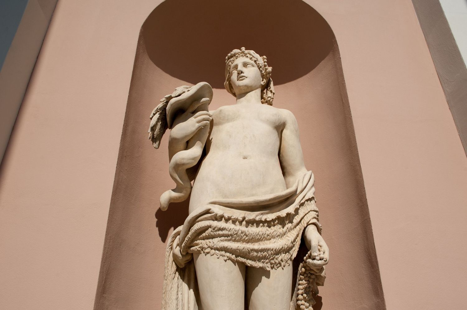 Classical marble statue of a semi-draped figure displayed in an architectural niche with pink walls.
