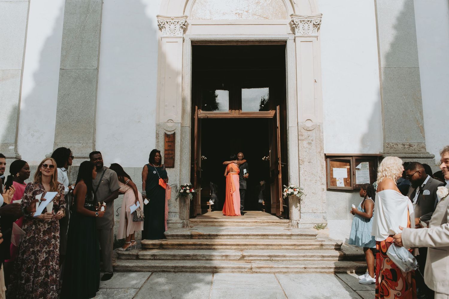 Wedding guests gather outside a white church as a bride in a pink gown descends the steps.