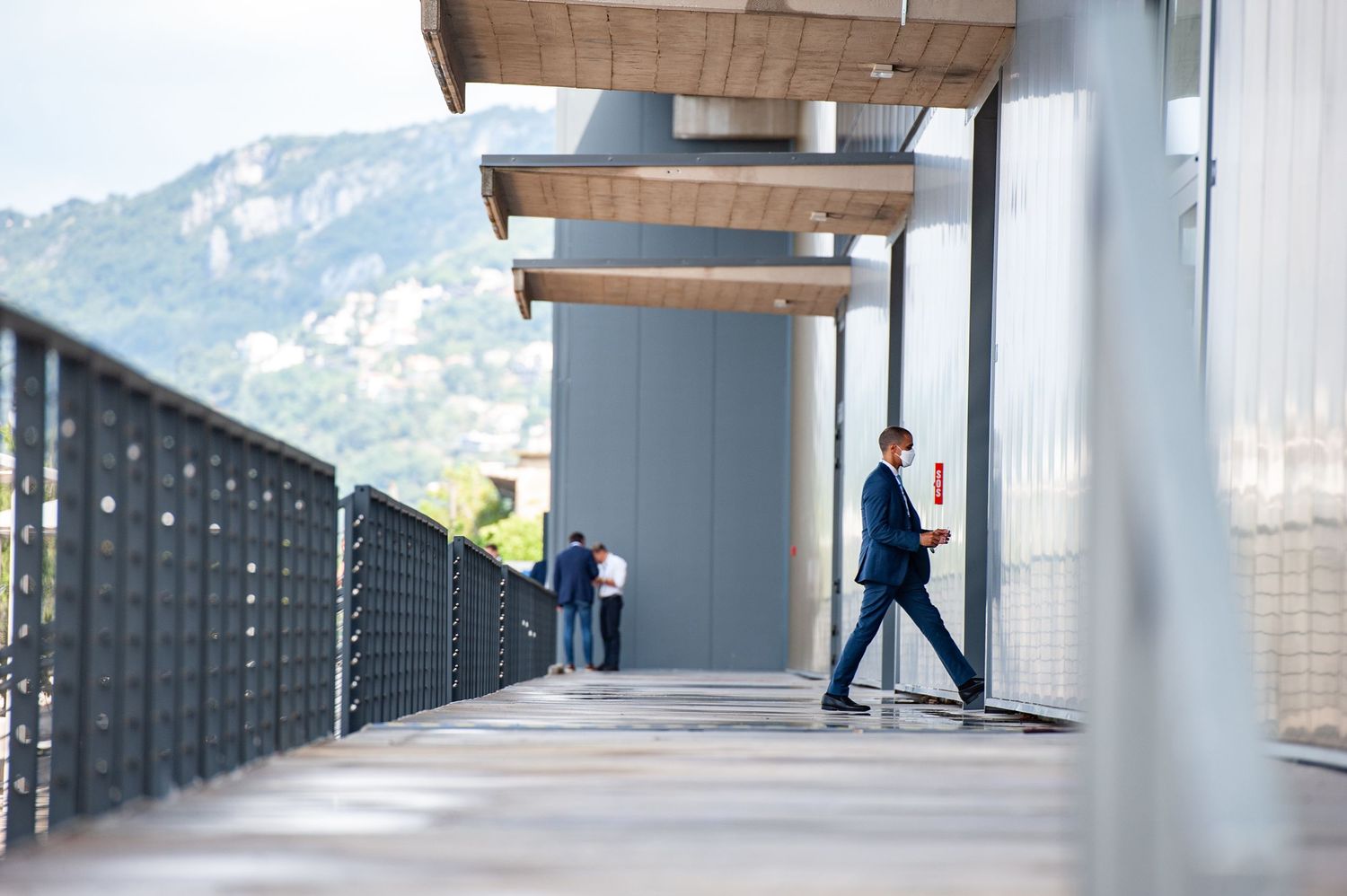 Businessman in navy suit walking on modern elevated walkway with mountain views and architectural columns.