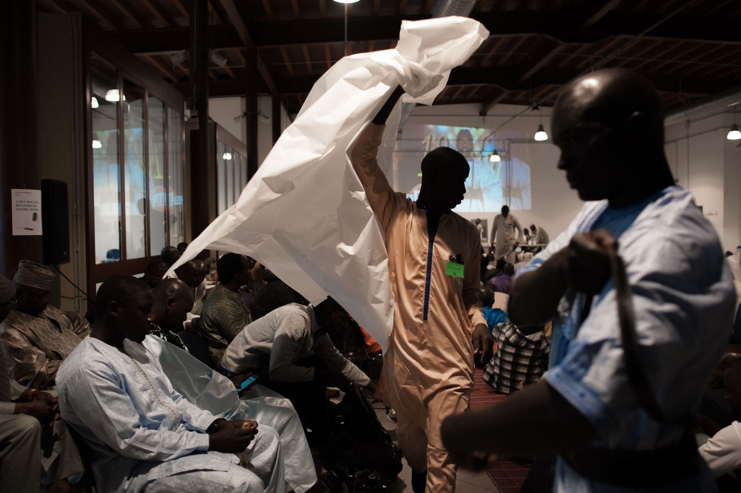 People gather indoors as a man waves a white cloth above a crowd in what appears to be a community meeting in Africa.
