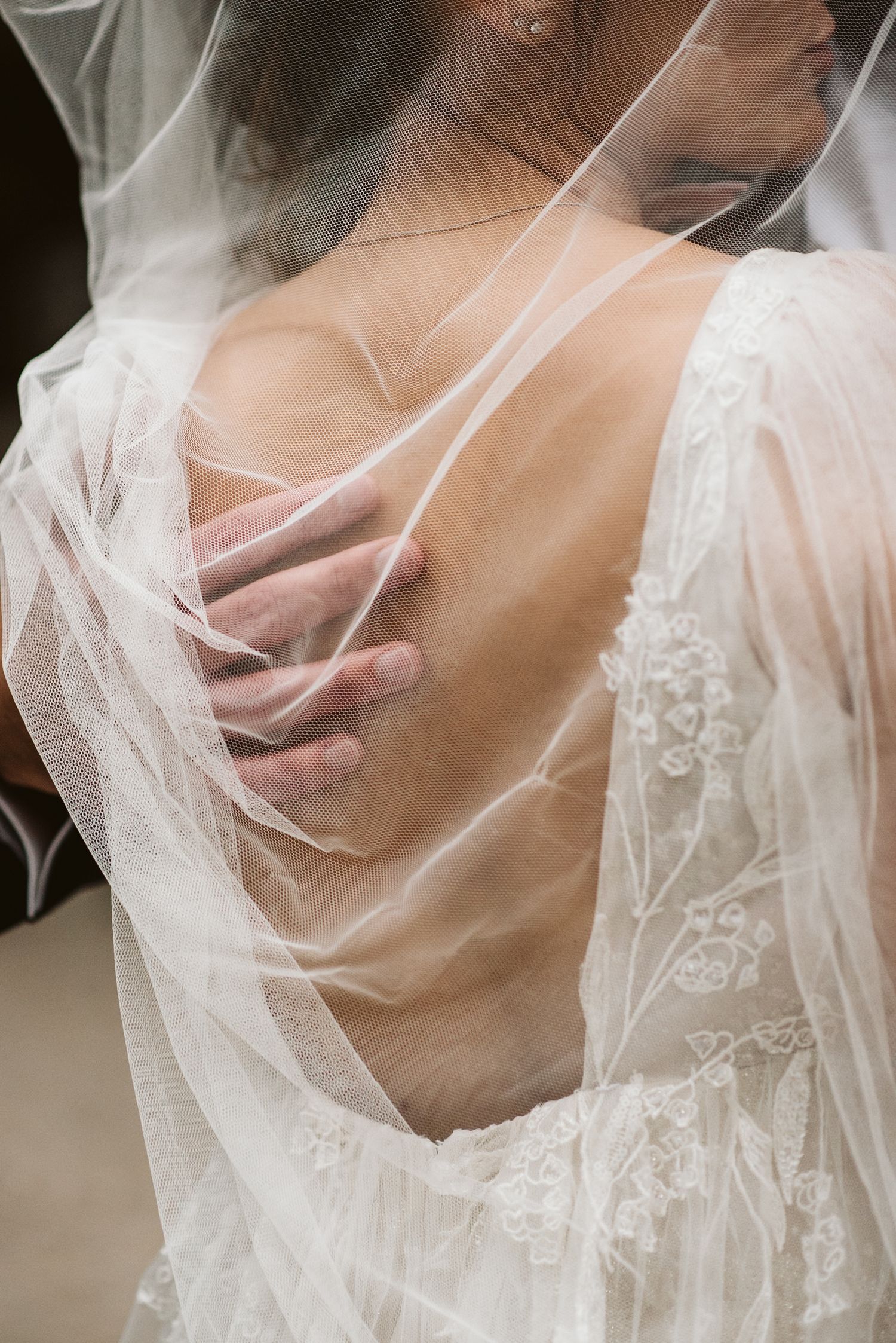 Bride in lace wedding dress with sheer veil draped over her back, groom's hand resting on her spine.