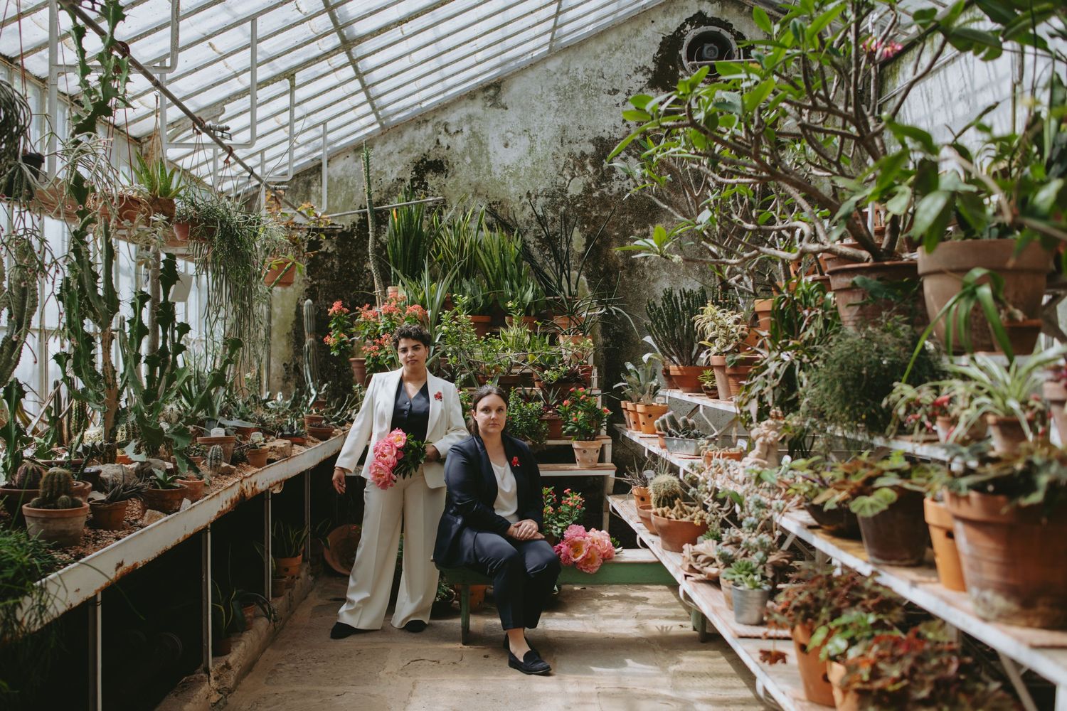 Two brides pose among lush potted plants and cacti inside a sunlit vintage greenhouse.