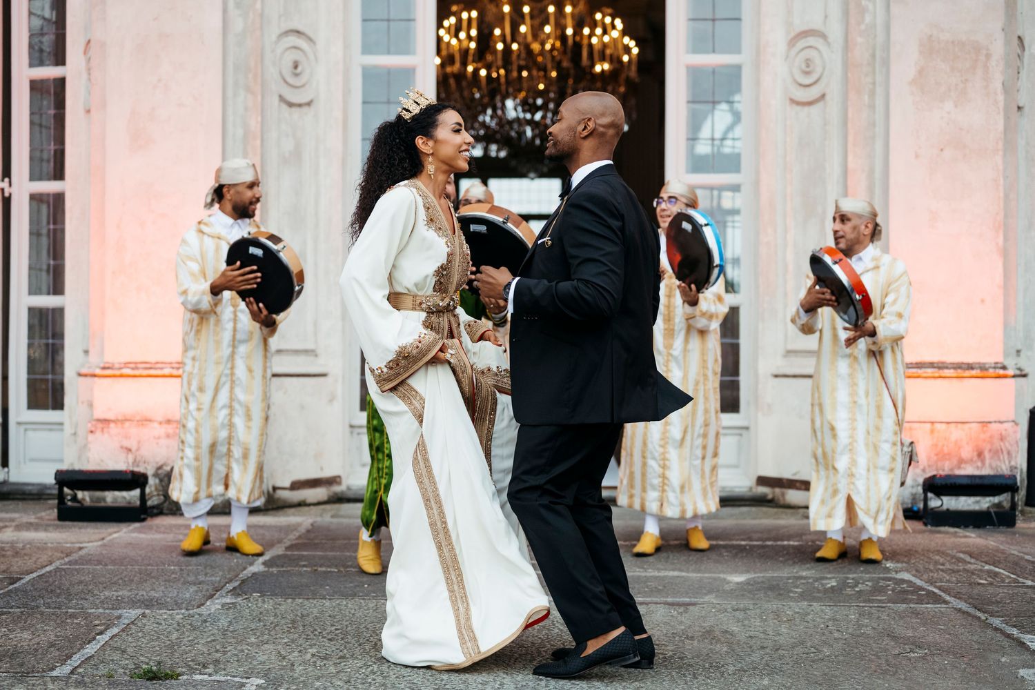 Couple in traditional Moroccan wedding attire dancing outdoors, with musicians in colorful robes performing in the background.
