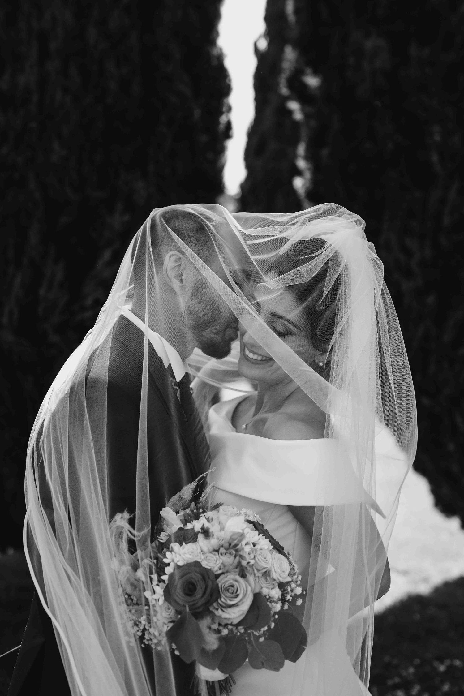Black and white photo of bride and groom sharing a romantic moment under a flowing wedding veil, bride holding a bouquet.