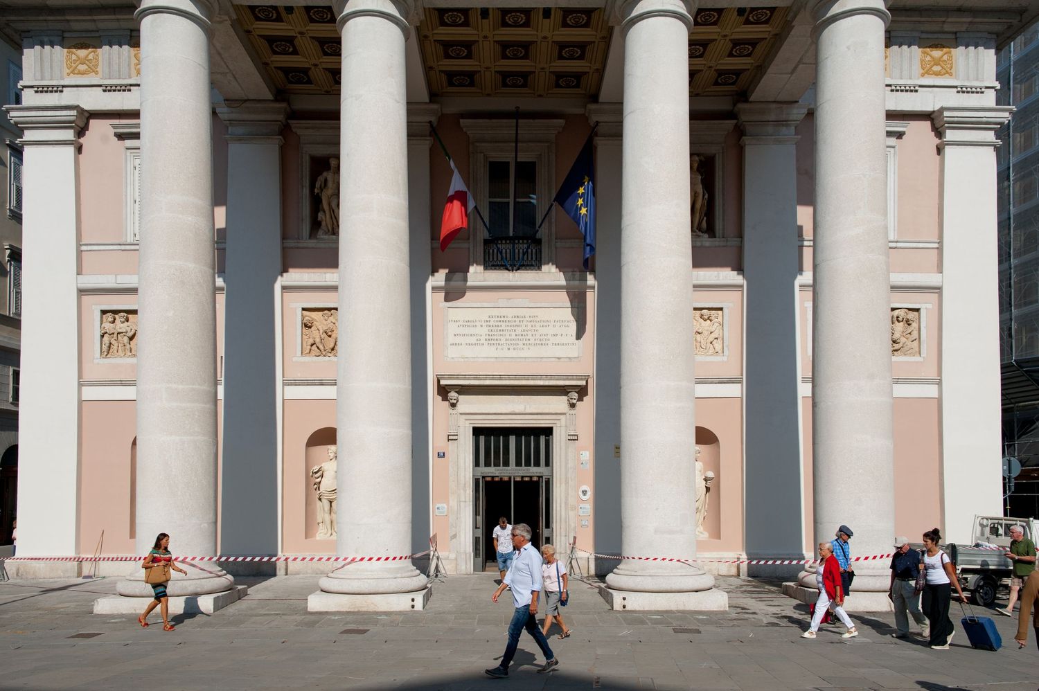 Neoclassical building facade with tall columns, flags, and people walking in front on a sunny day.