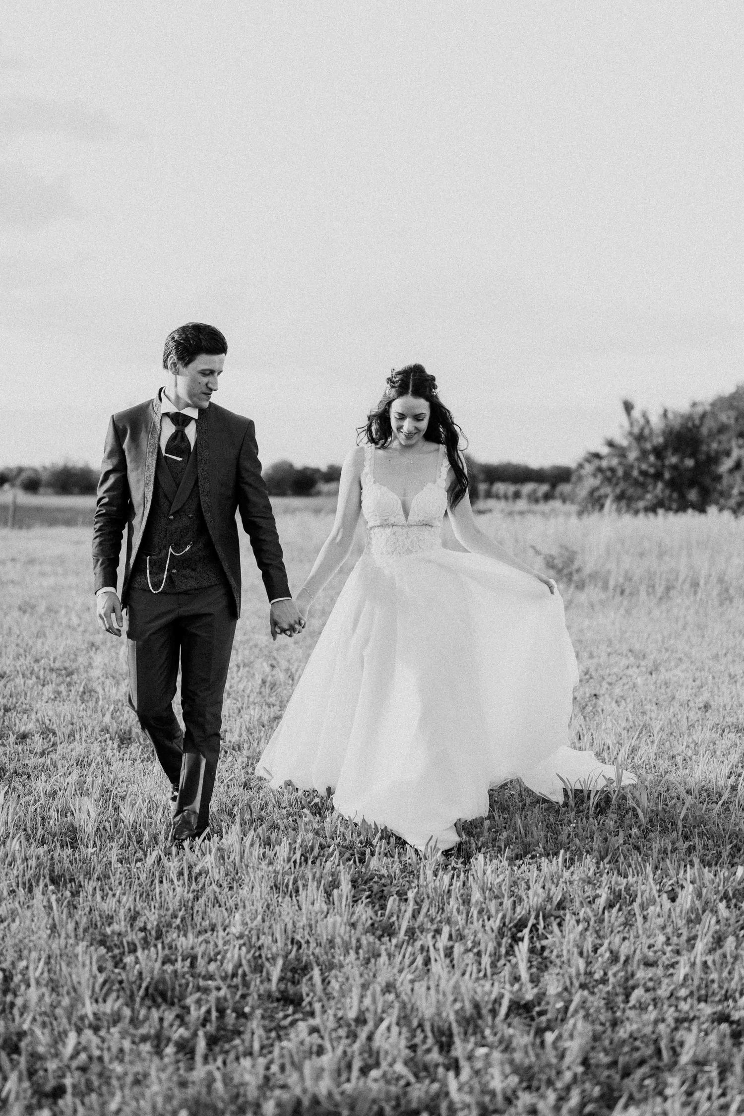Black and white photo of a bride and groom walking hand in hand through a field on their wedding day.