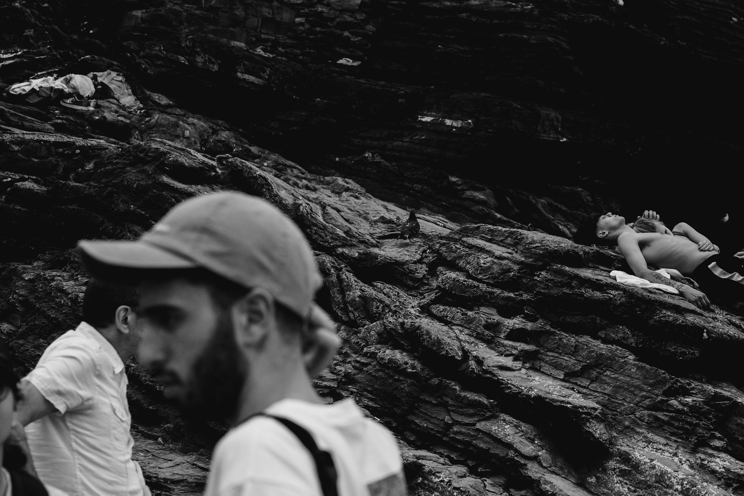 Black and white photo of bearded man in cap with person resting on rocky terrain in background.