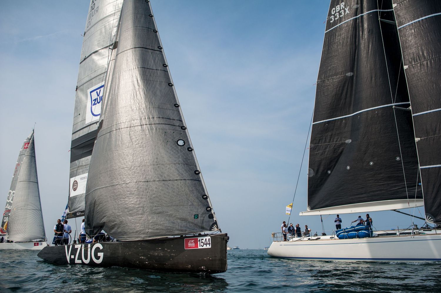 Two racing sailboats with dark sails compete on open water during a regatta, with V-ZUG visible on the lead vessel.