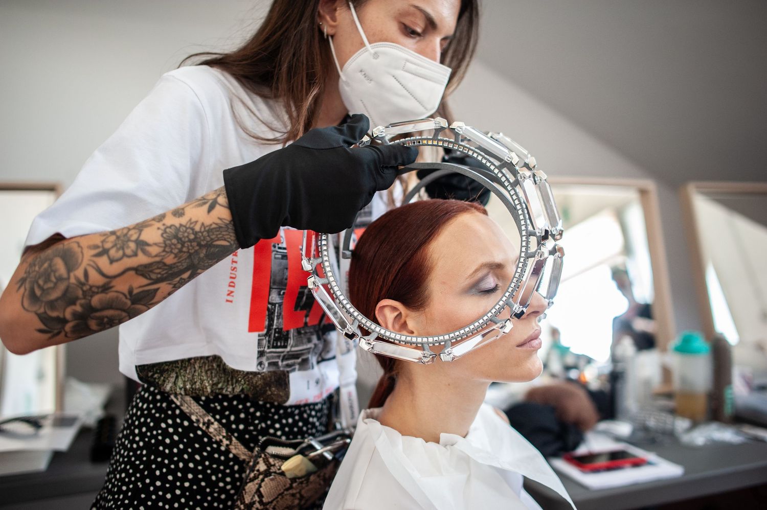 Tattooed hairstylist wearing mask styles redhead client's hair using decorative mirror in bright salon.