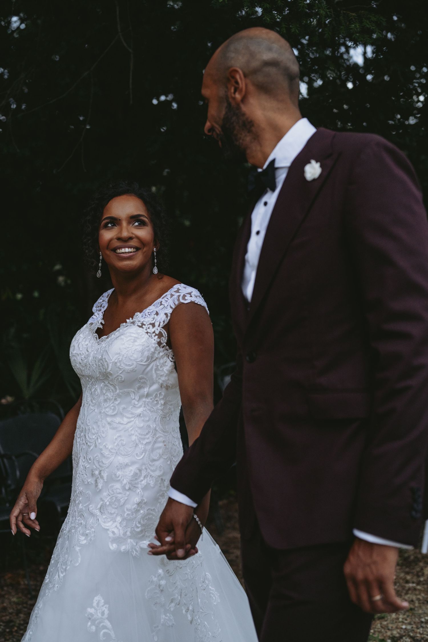 A bride in lace gown and groom in burgundy tuxedo hold hands and smile during their outdoor wedding ceremony.