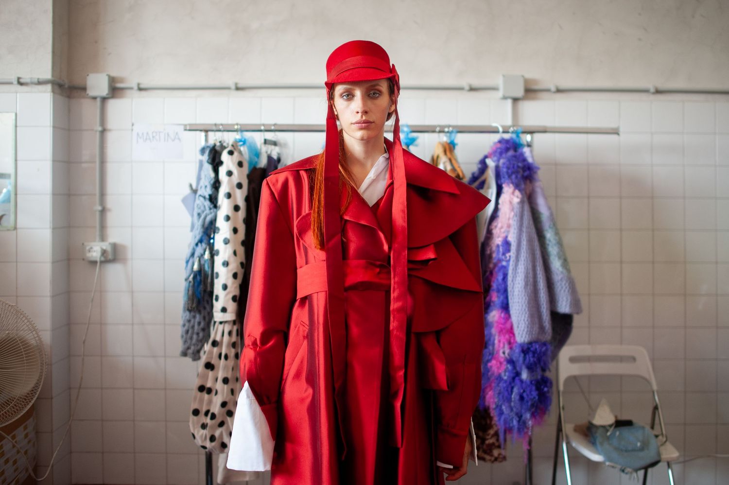 Woman in red coat and cap poses in front of clothing rack in tiled room.