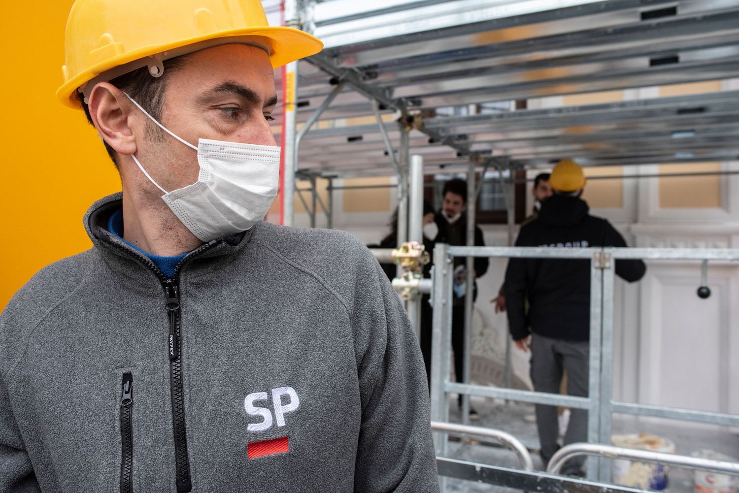 Construction worker wearing yellow hard hat and face mask at building site with colleagues working in background.