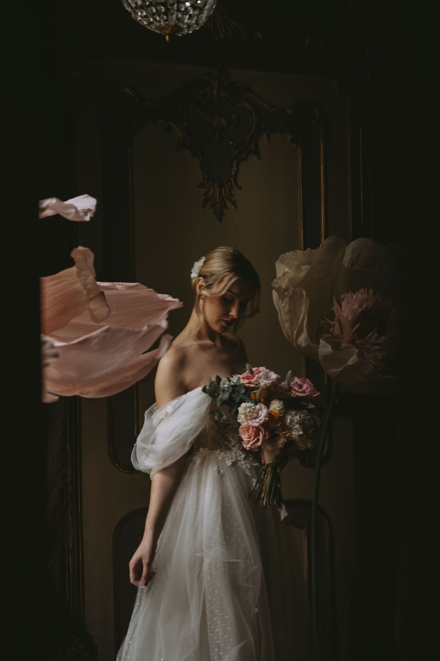 A person in a flowing white wedding dress holds a romantic bouquet of pink and red flowers in a dark moody setting.