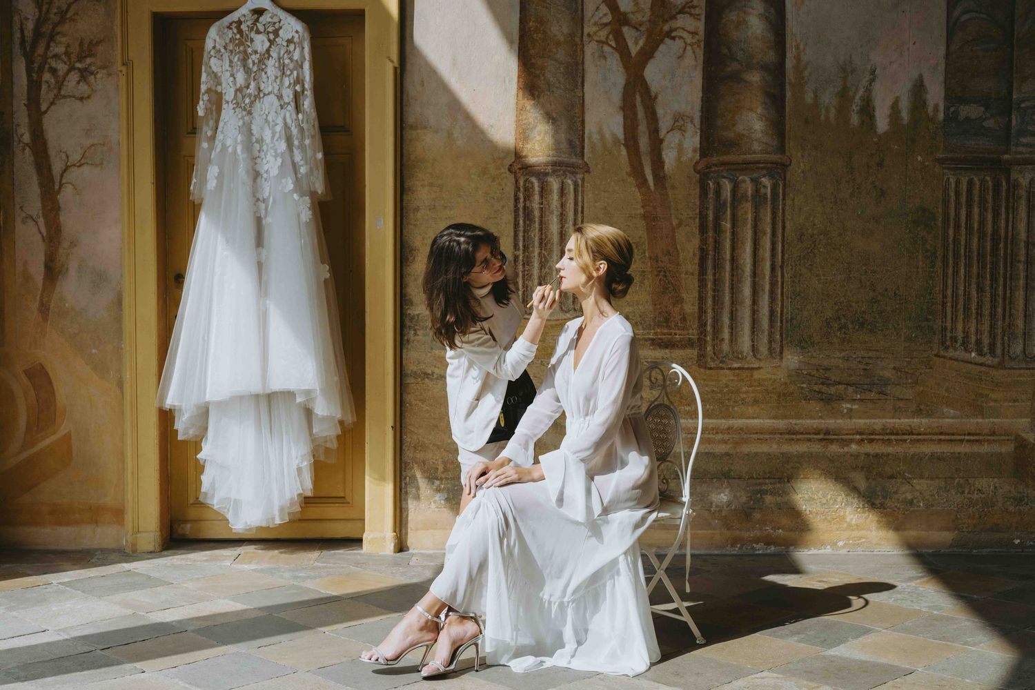 Bride getting makeup done by stylist in elegant room with wedding dress hanging in sunlit background.