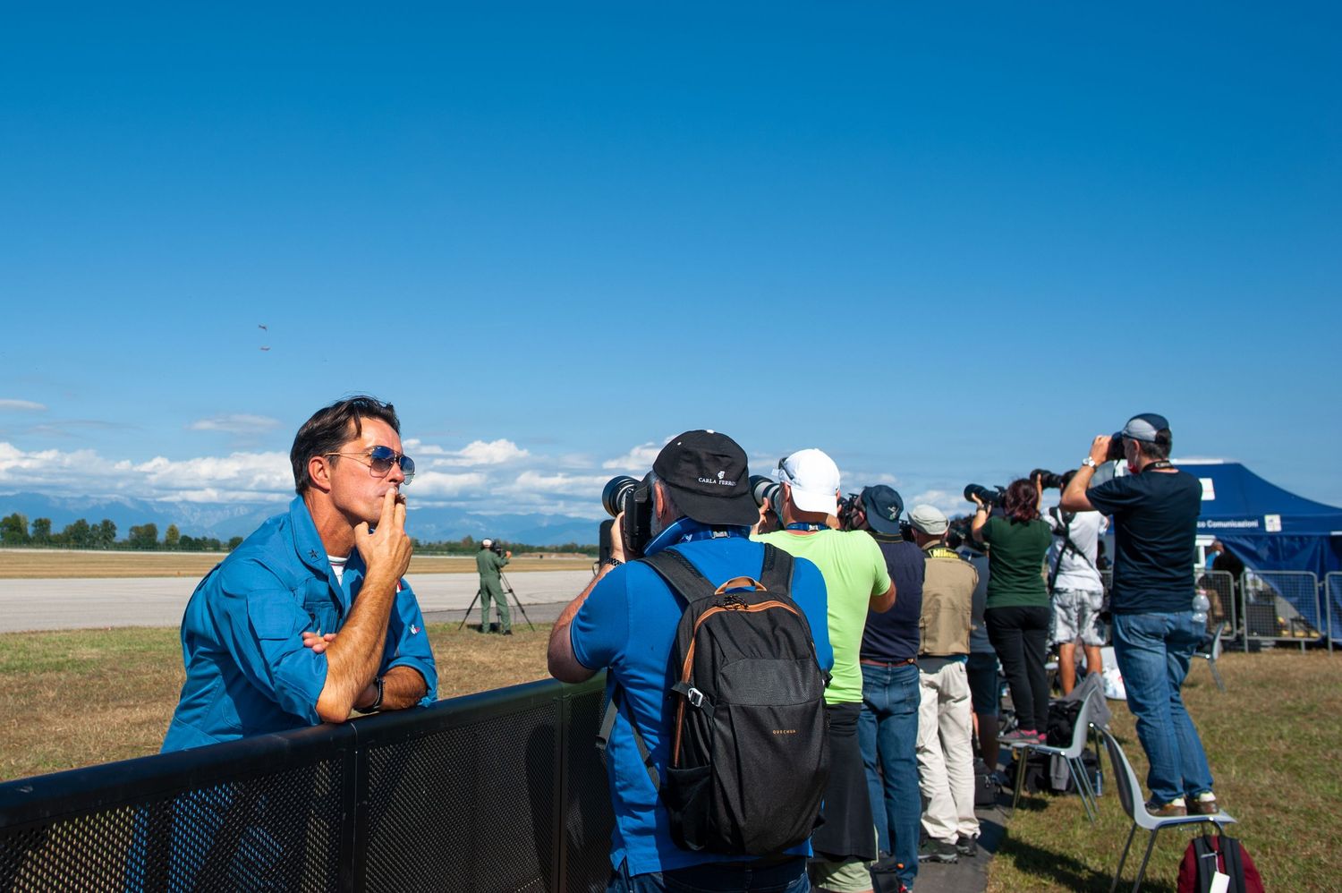 Spectators watch aircraft at an outdoor airshow on a sunny day near a runway with mountains in the background.