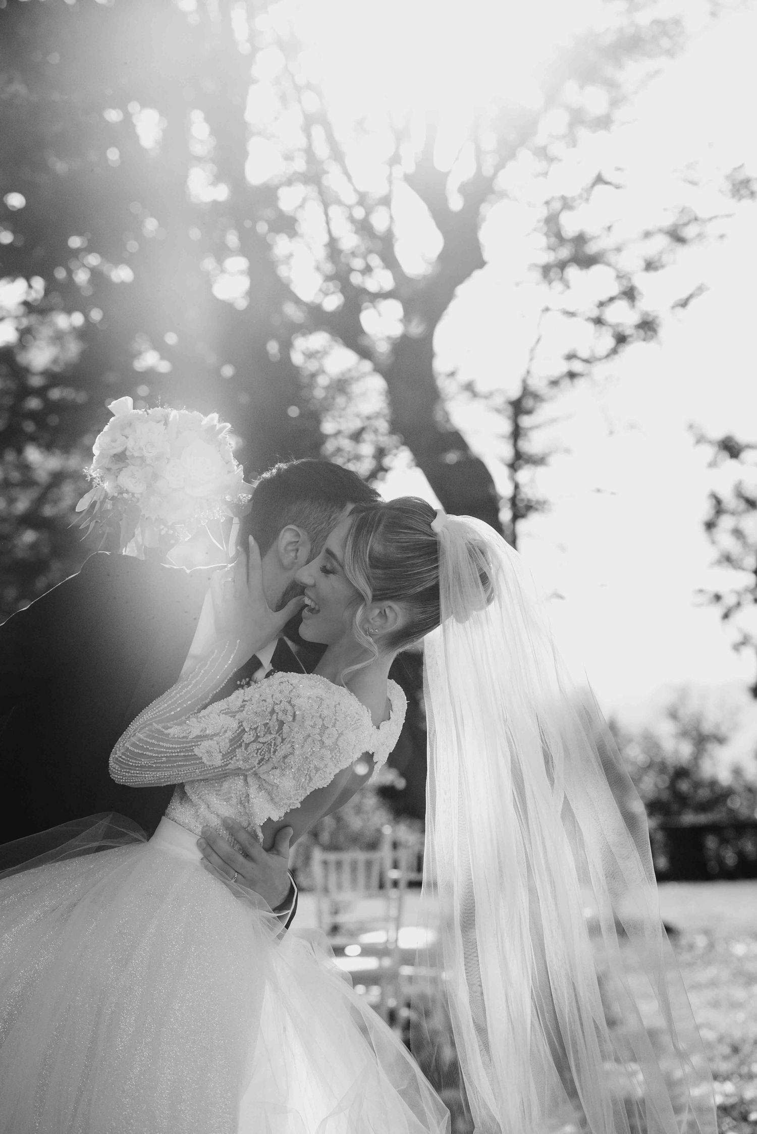 Black and white wedding photo of bride and groom kissing outdoors with sunlight and trees in background.