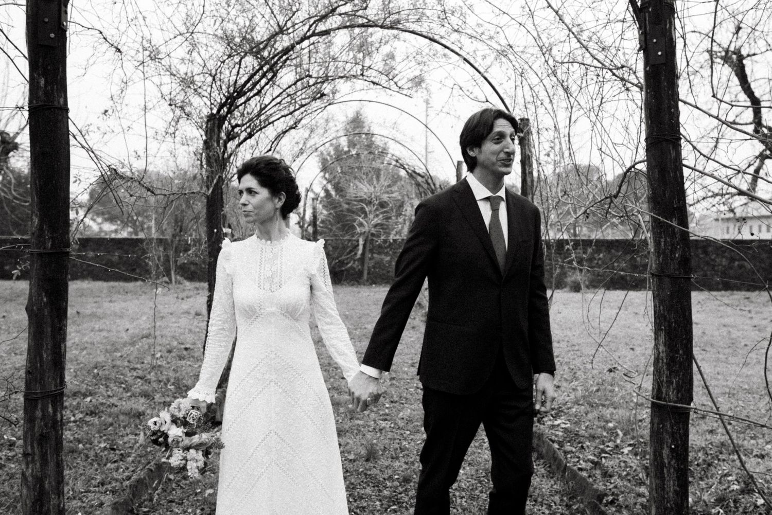 A bride and groom walk hand in hand through a forest of bare trees on their wedding day.