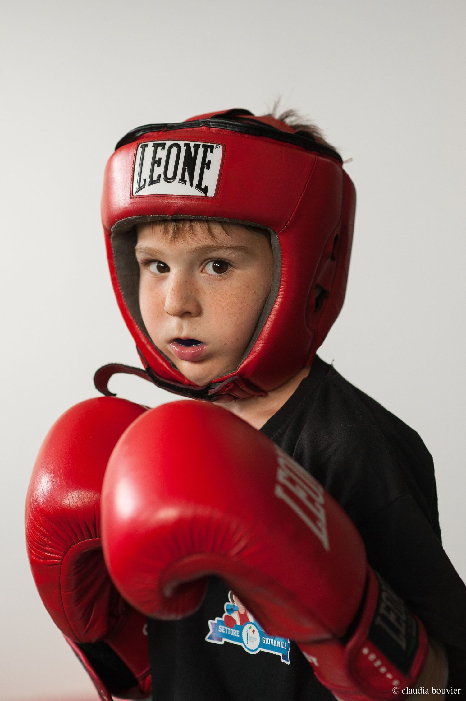 Young boy in red Leone boxing headgear and red gloves poses in fighting stance wearing black sportswear.