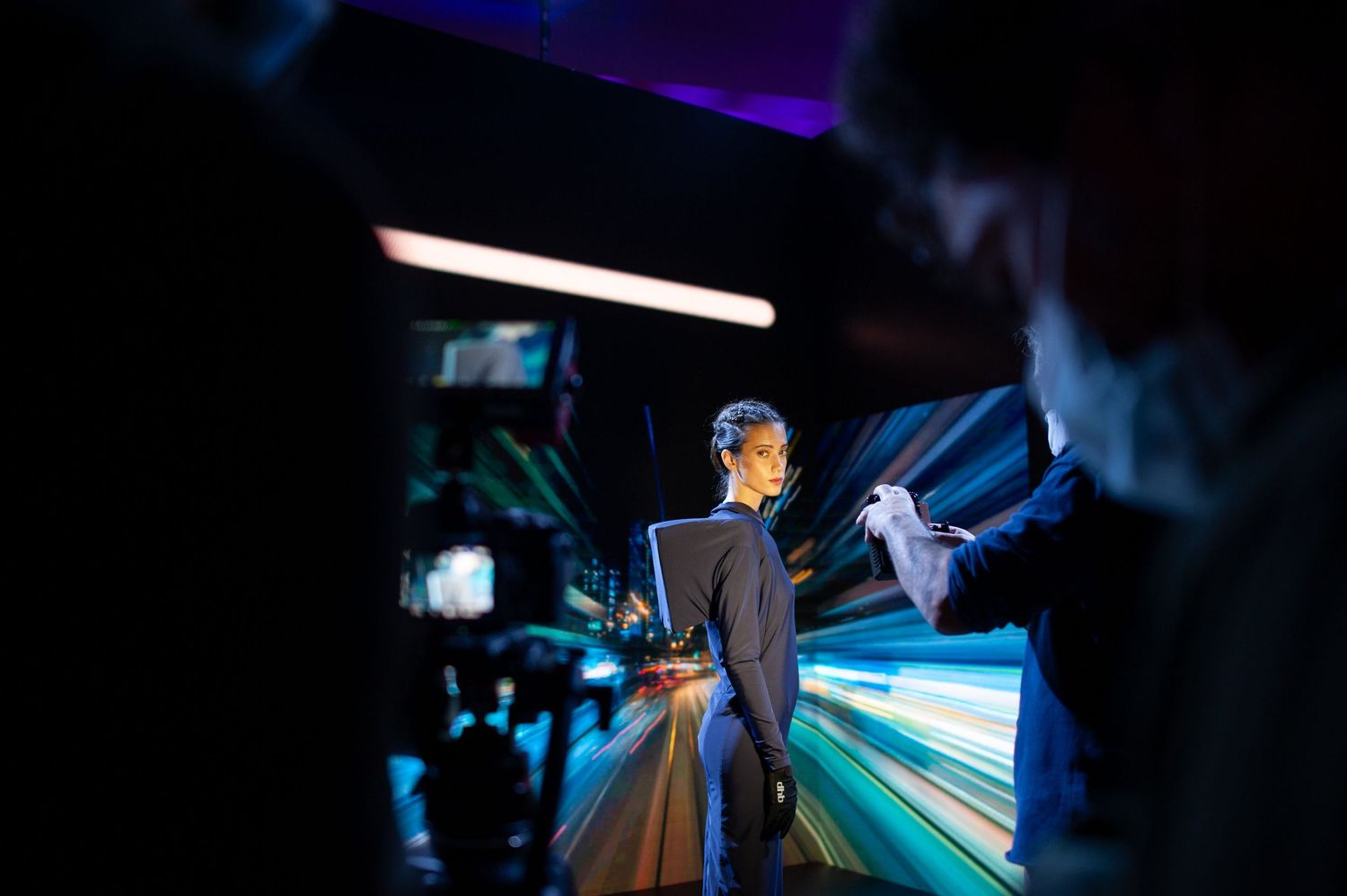 A presenter stands on a dramatic blue-lit stage during a professional event, surrounded by camera crew in the foreground.