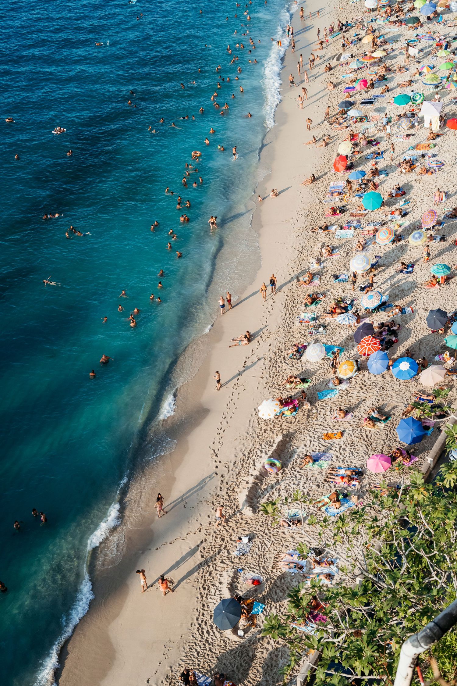 Aerial view of a crowded beach with colorful umbrellas, sunbathers on sand, and swimmers in turquoise ocean water.