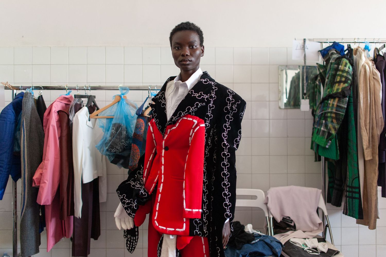 A model poses in a bold black and red embroidered jacket in a fashion backstage dressing room with clothing racks.