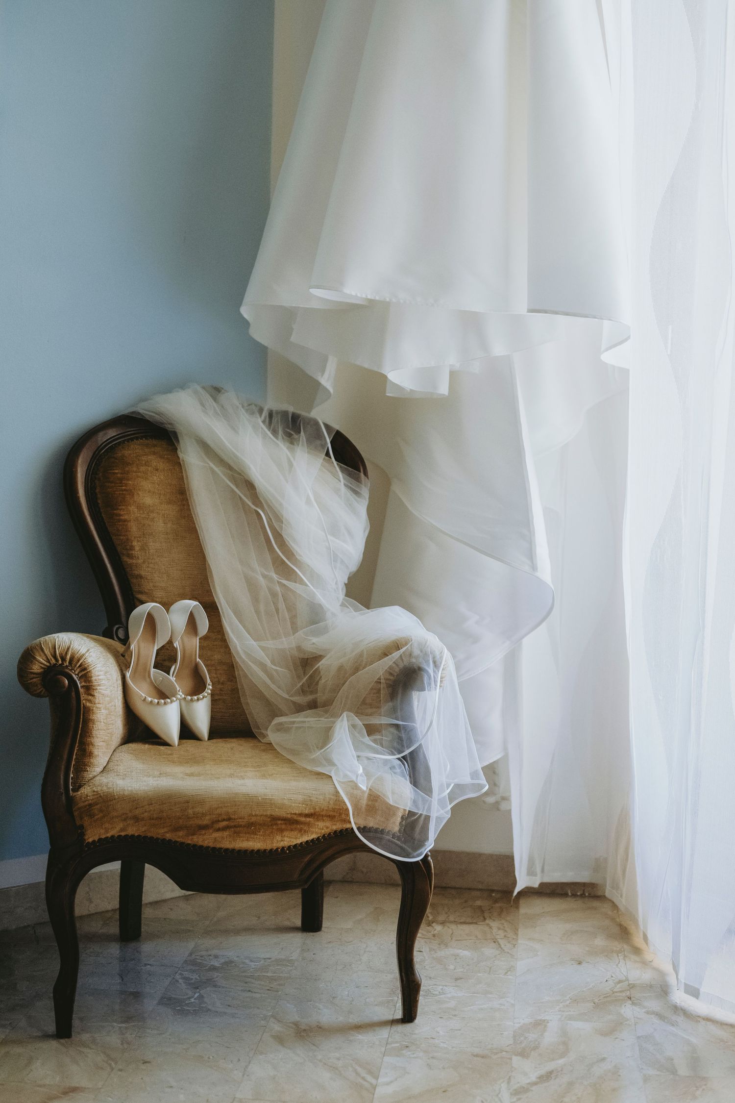 Elegant bridal veil and white heels on a vintage gold armchair beside a flowing wedding dress near a curtained window.